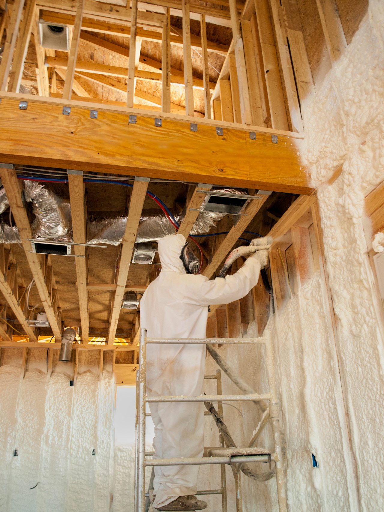 A worker in a white suit and respirator applies spray foam insulation in a residential building under construction, focusing on energy efficiency and thermal protection.